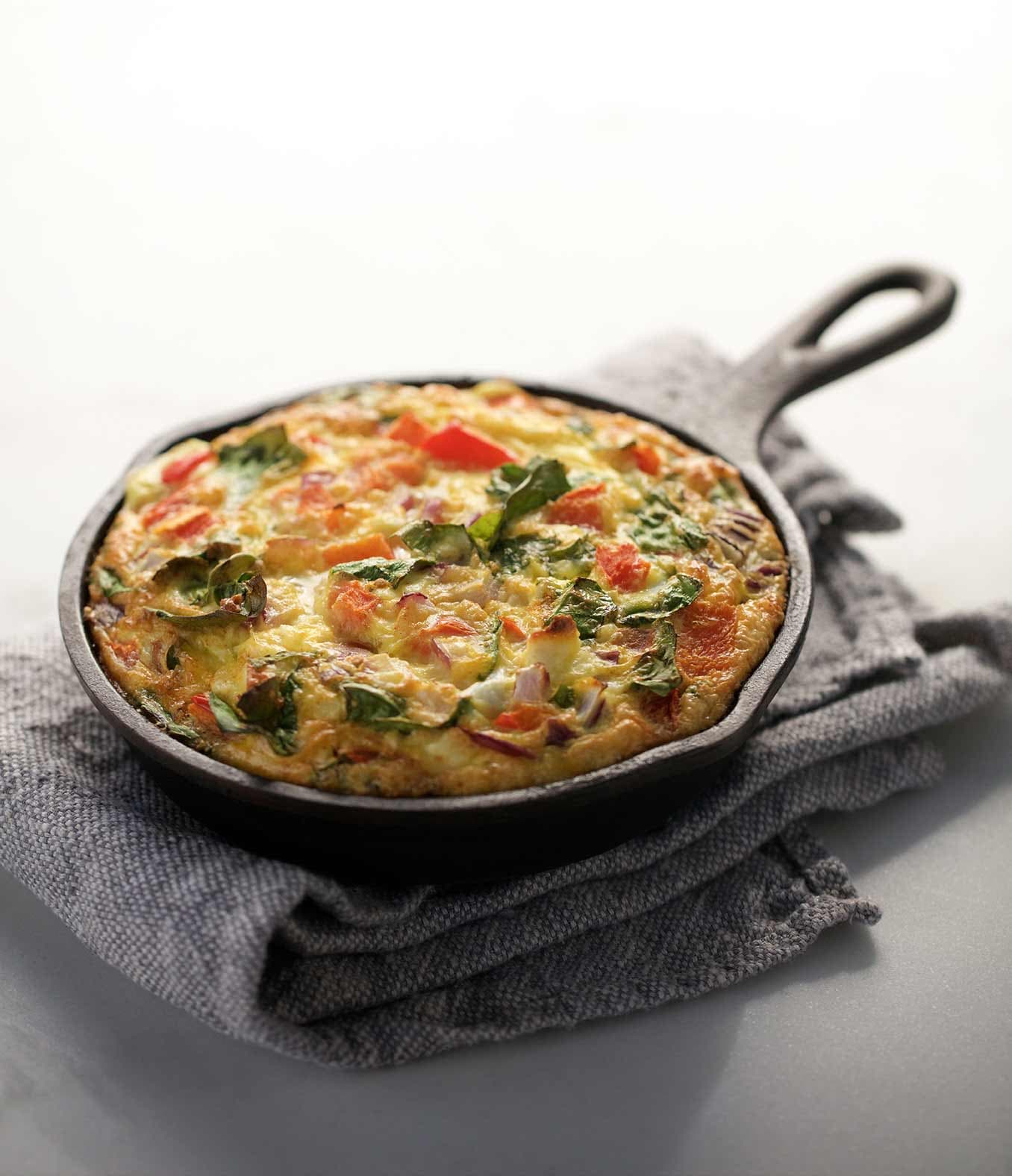 Frittata with onion, peppers, spinach and tomatoes in a cast iron skillet resting on a towel.