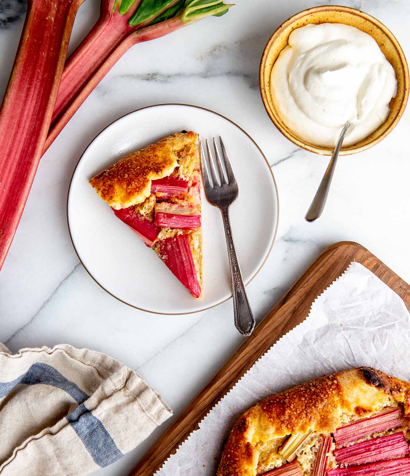 Slice of rhubarb pie on a plate with a fork and a side bowl of whipped topping.